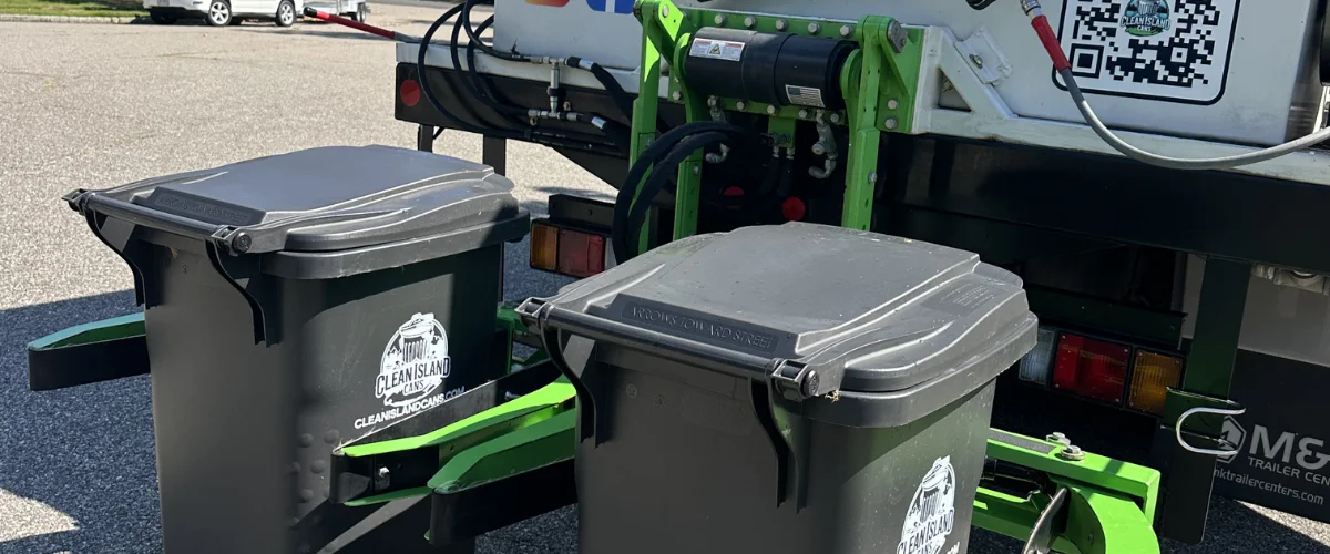 Two men standing next to a trash can cleaning truck, with a rainbow visible in the spray.