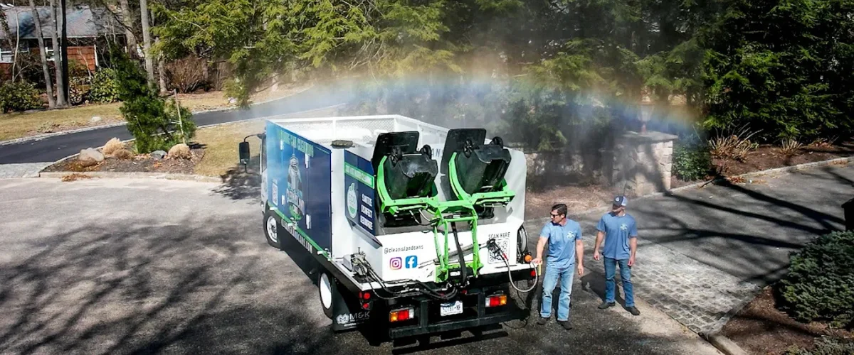 Two men standing next to a trash can cleaning truck, with a rainbow visible in the spray.