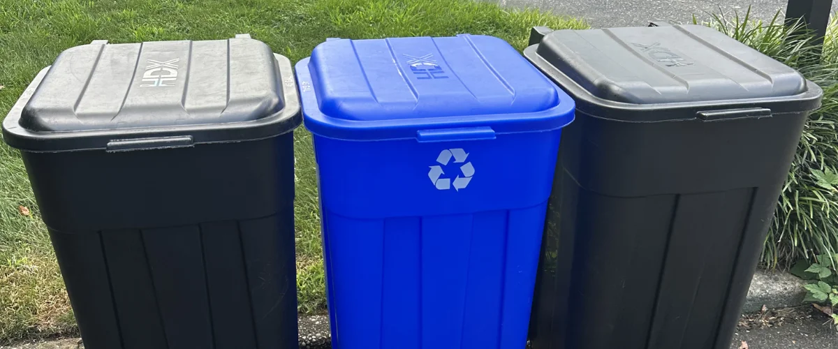 Three bins, two black and one blue with a recycling symbol, stand side-by-side on green grass. Three bins, two black and one blue with a recycling symbol, stand side-by-side on green grass.