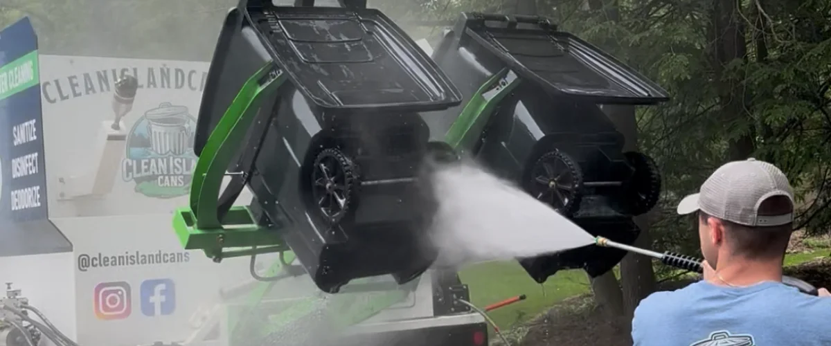 A man in a cap power washes two large black trash cans, tilted on a white truck with green accents. A man in a cap power washes two large black trash cans, tilted on a white truck with green accents.