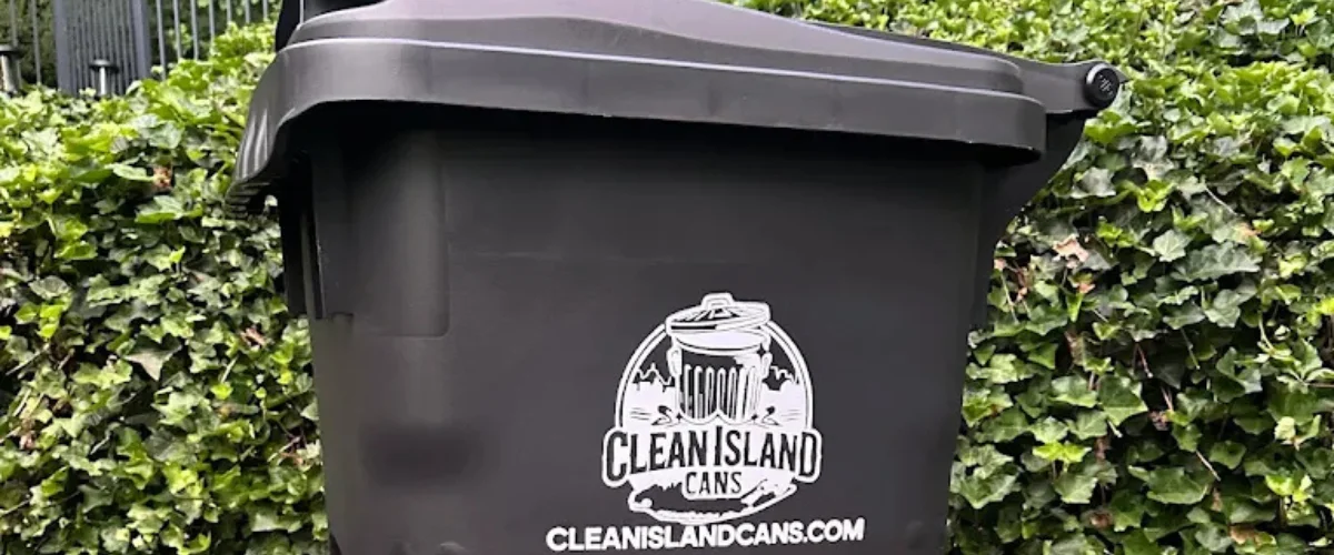 A black plastic waste bin with the white 'Clean Island Cans' logo, set against a backdrop of green ivy.