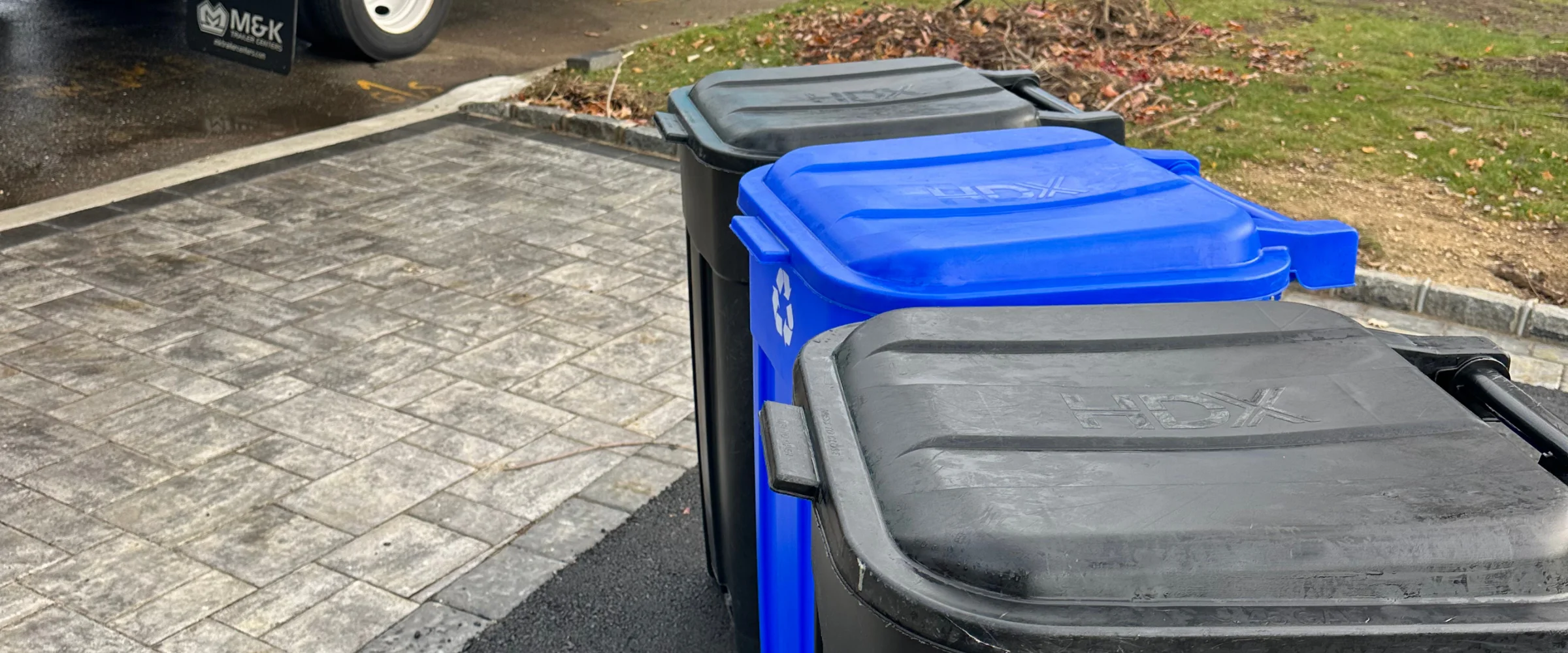 Three outdoor bins, one blue recycling and two black, lined up on a grey paved driveway with grass nearby.