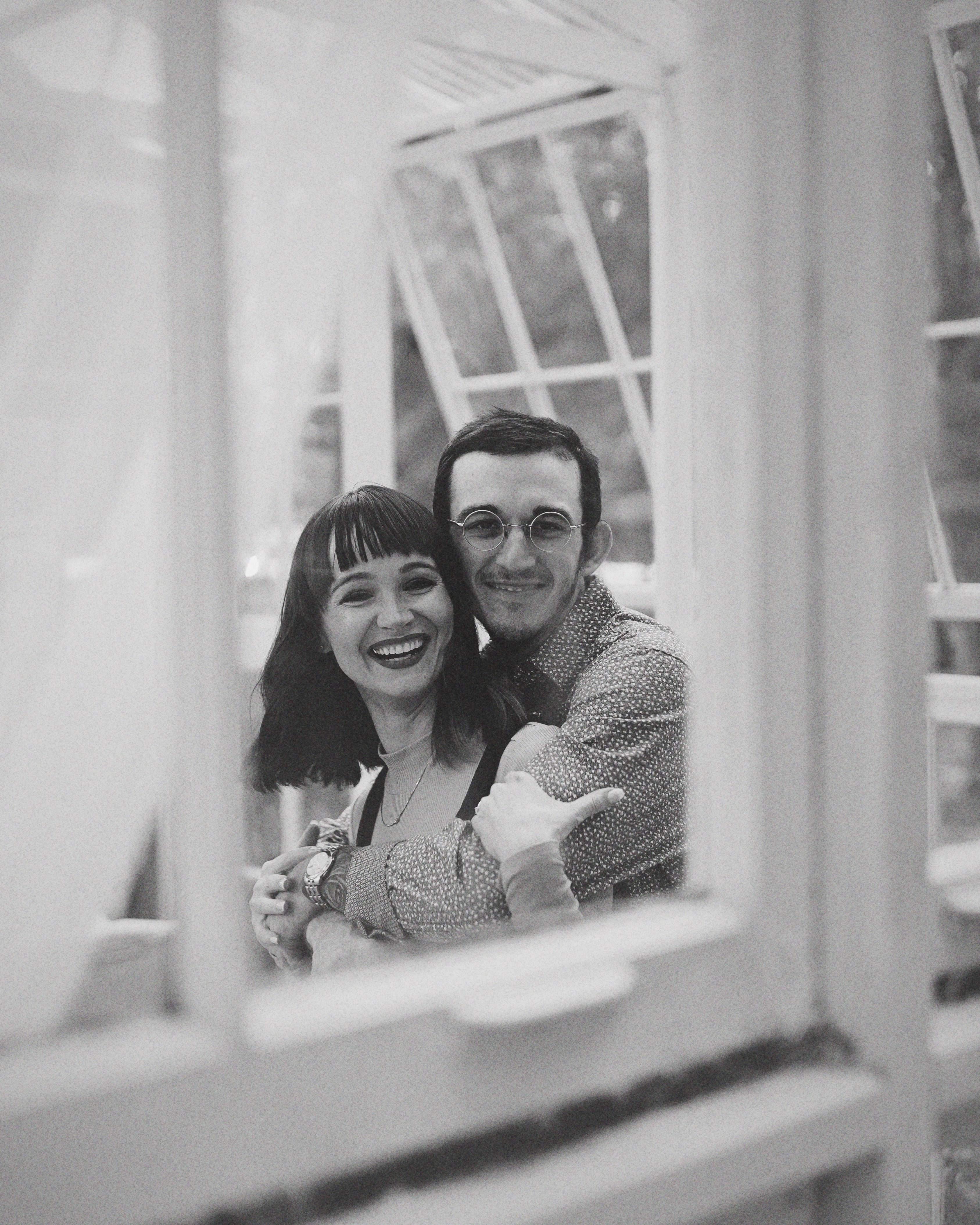 Image of an engaged couple laughing at the camera through the window of The Cottage Greenhouse in Kilgore, Texas.