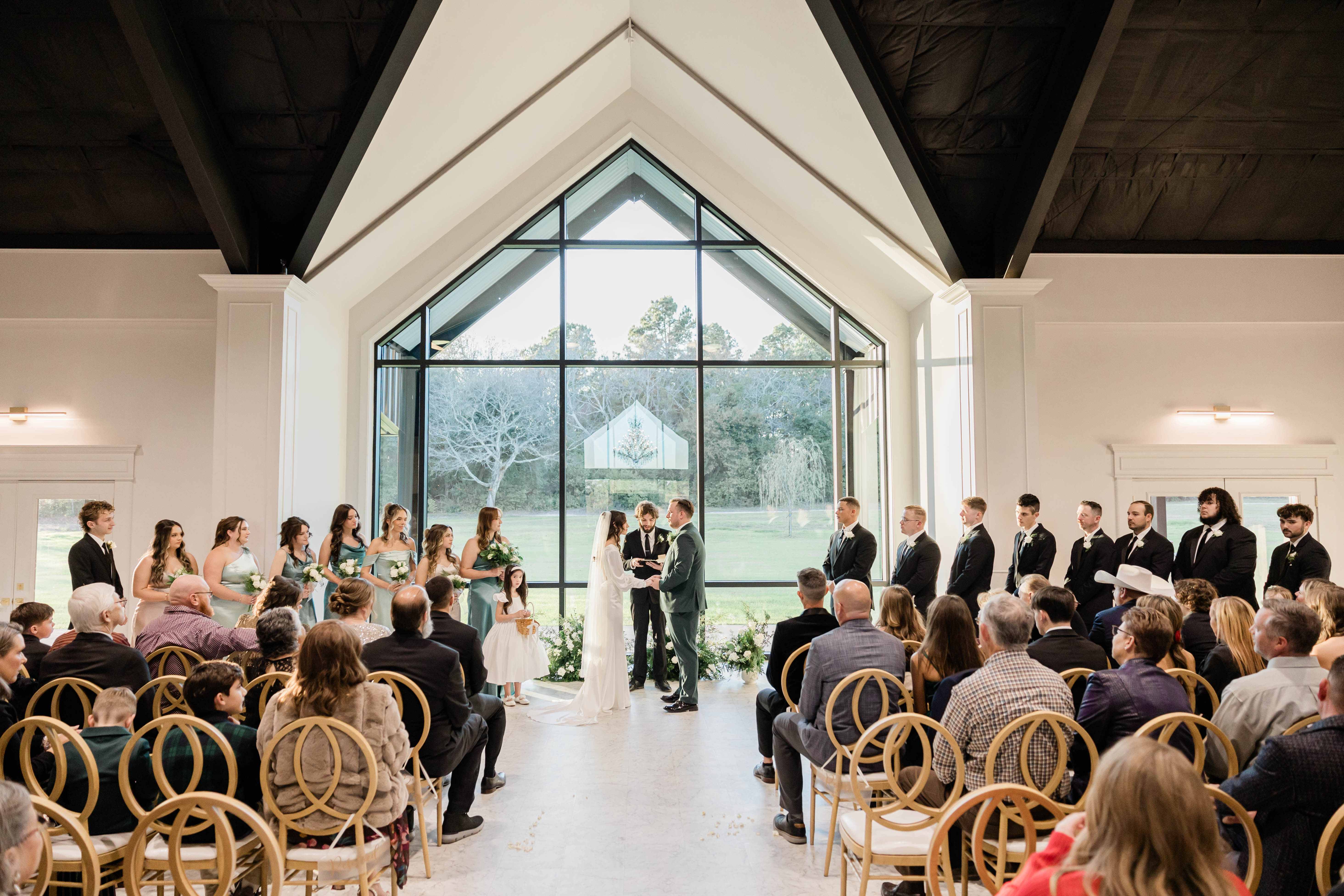 A picture of the wedding ceremony from behind the guests showing off the beautiful cathedral window backdrop behind the couple at Briar Rose Venue in Tyler, Texas.