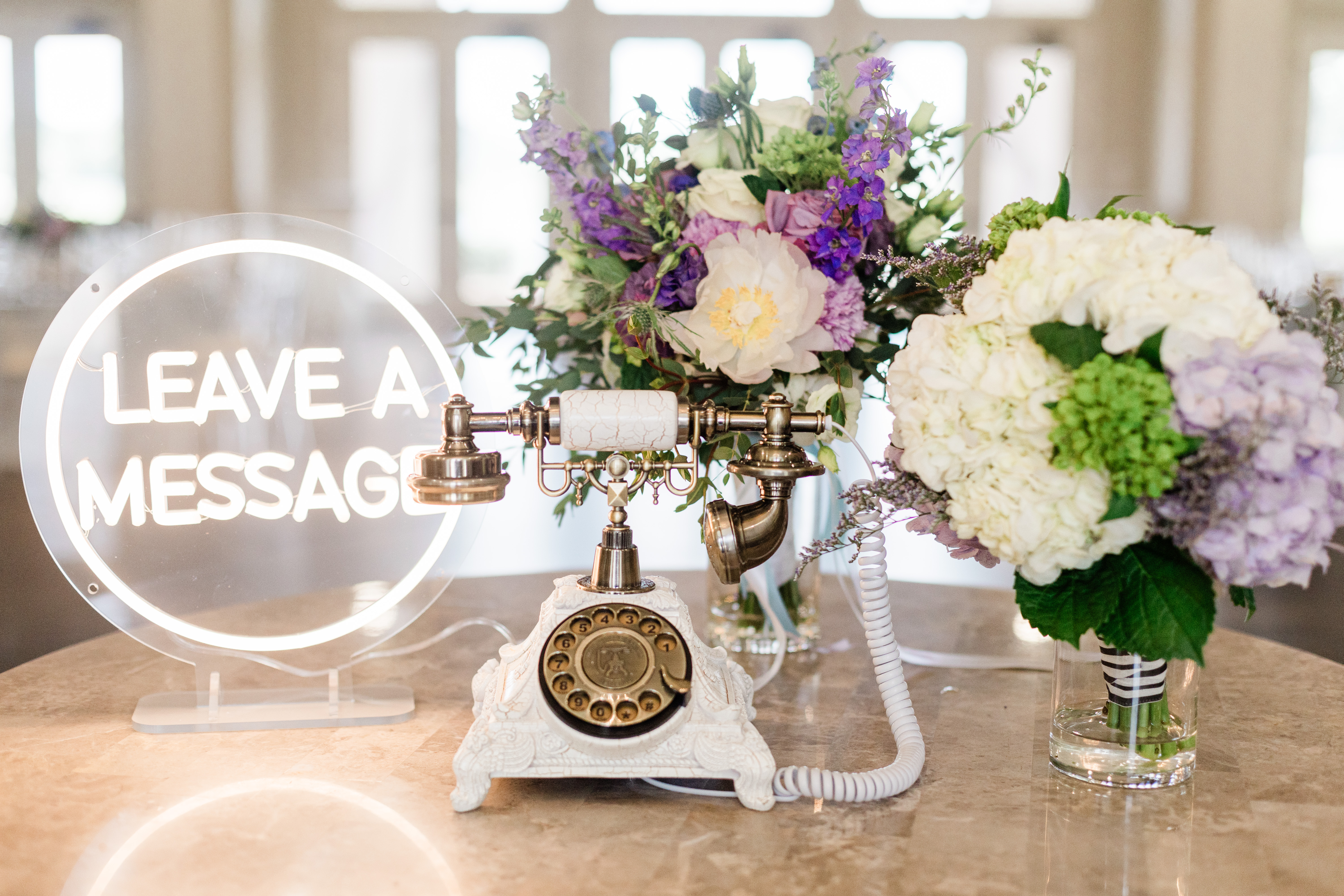 A picture of an antique white phone with two floral arrangements and an acrylic neon "leave a message" sign.