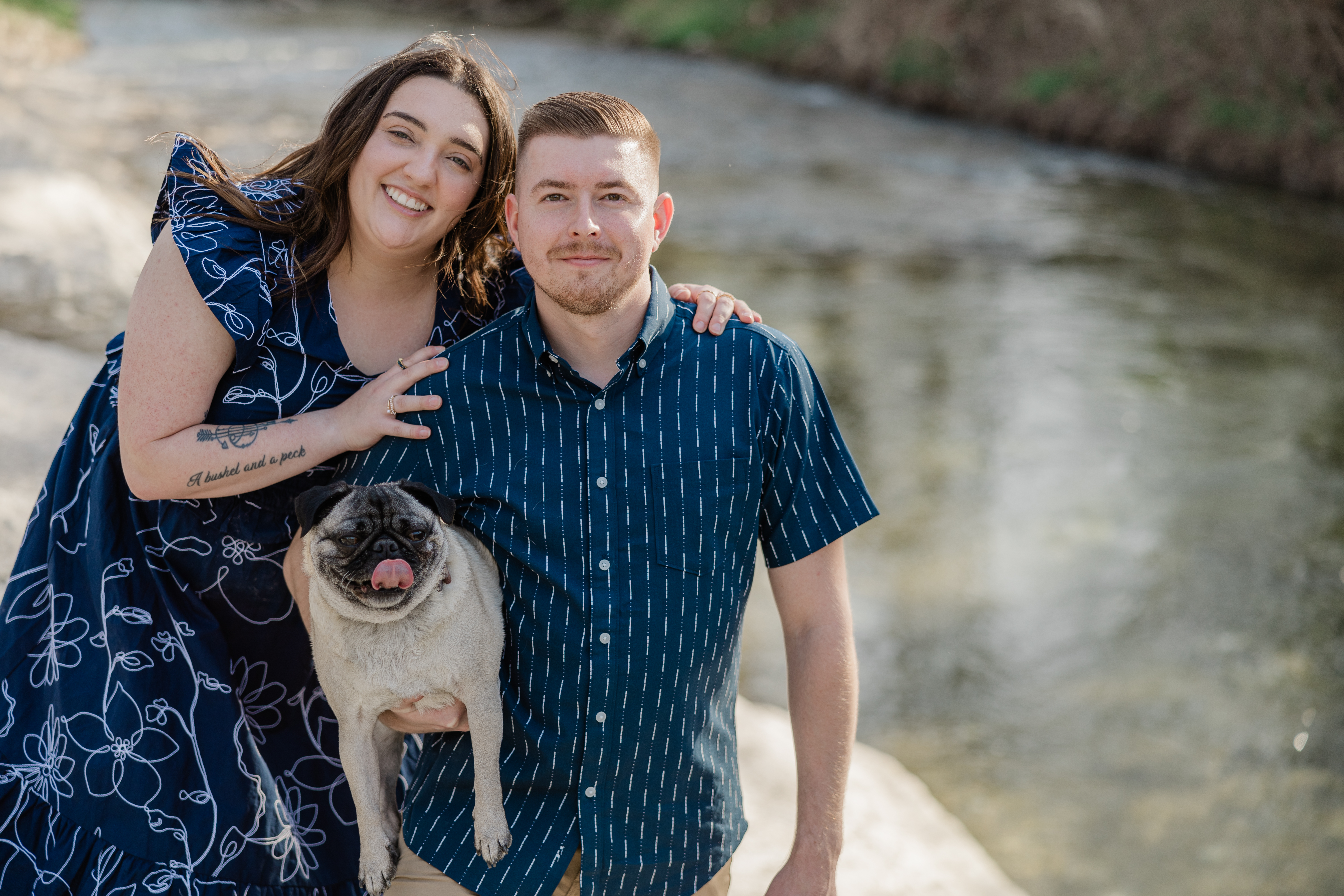 Image of a couple with their Pug named Higgins having their engagement photos taken at a park in Richardson with a flowing creek in the background captured by proposal and engagement photographer in Dallas, Arlene Stepanian.