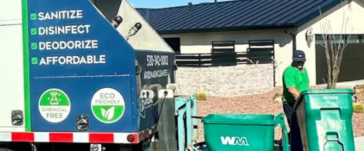 A service worker in a green shirt and hat, standing beside a large green waste bin, with a blue truck behind him.