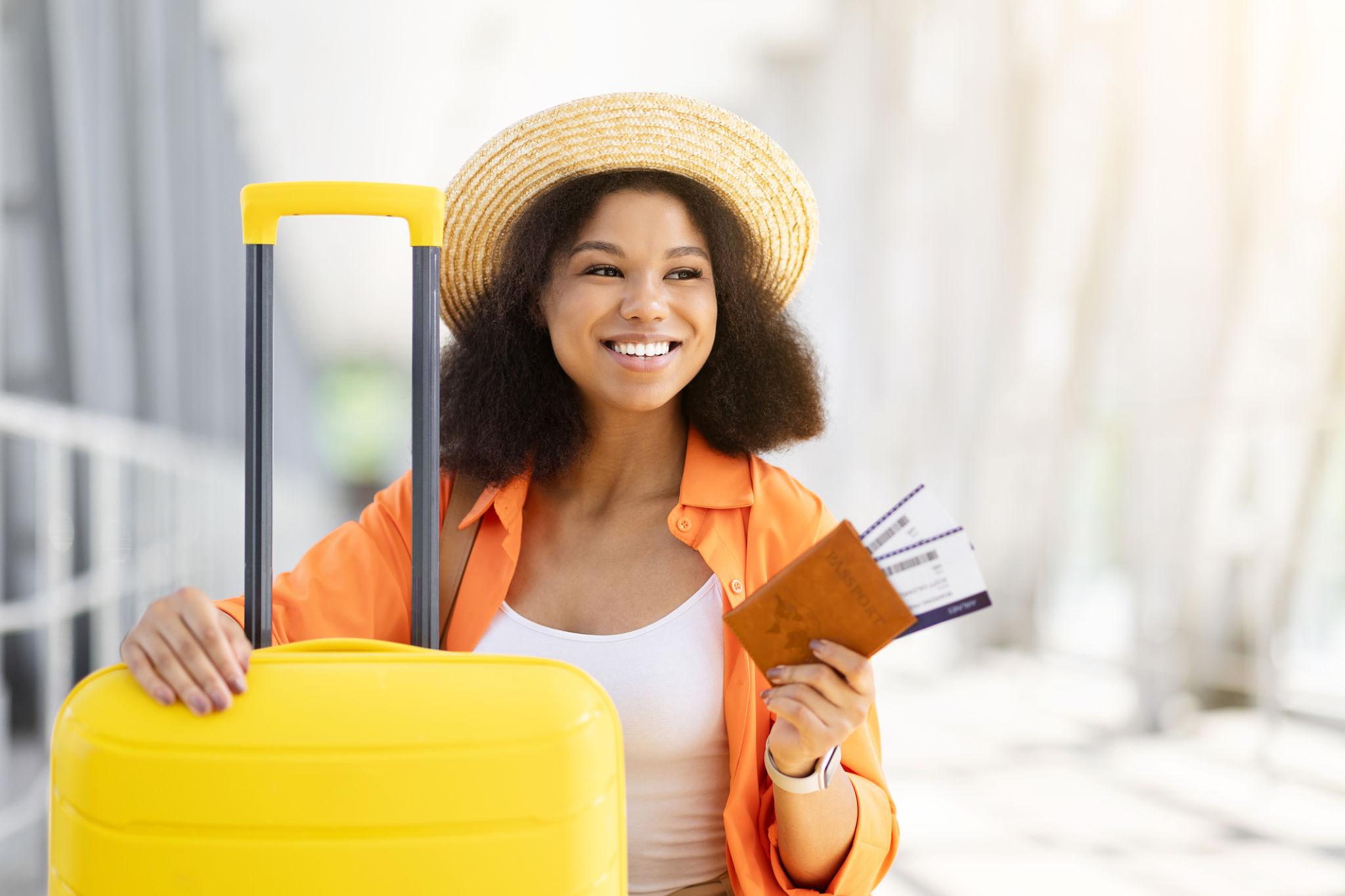 Black woman traveler sitting beside suitcase at airport, holding passport and tickets