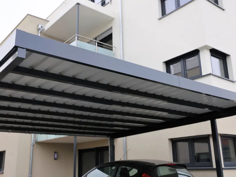 Modern flat-roof carport with slatted metal panels covering a parked car. Modern flat-roof carport with slatted metal panels covering a parked car.