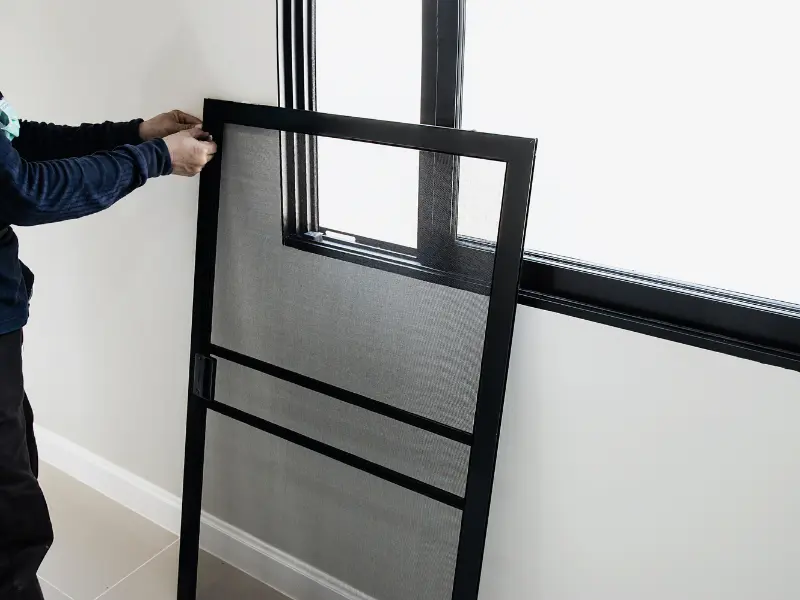 Installer positioning a framed mesh screen panel against a window opening during screen room installation.