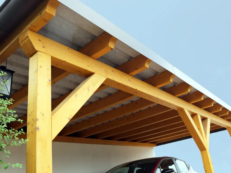 Wood-frame carport with exposed beam roof over a driveway.