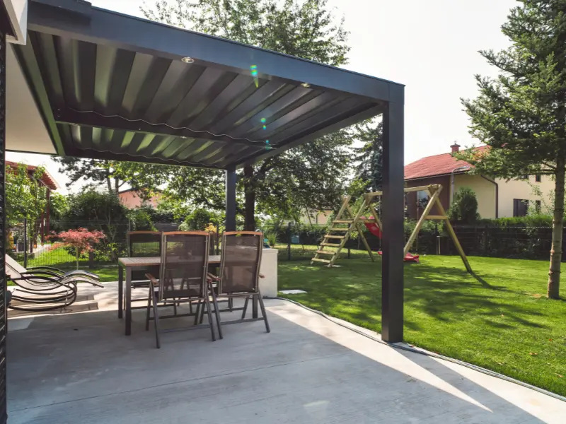 Dark metal solid patio cover with built-in lights shading an outdoor dining table and lounge chairs beside a green lawn and play set.. Dark metal solid patio cover with built-in lights shading an outdoor dining table and lounge chairs beside a green lawn and play set..