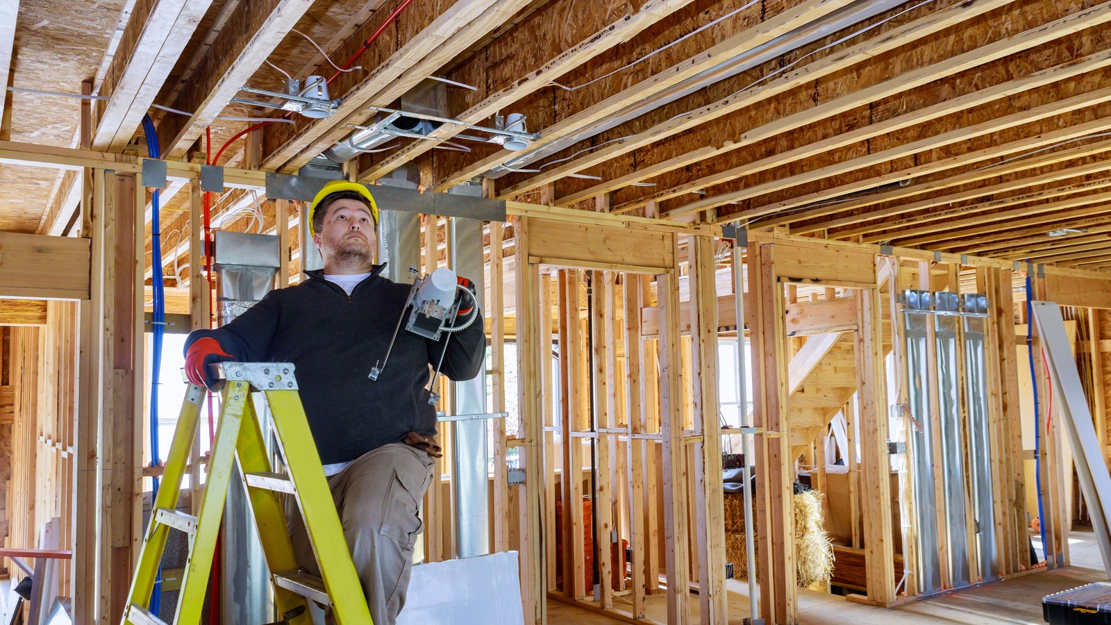 An electrician on a ladder working on ceiling wiring in a framed interior