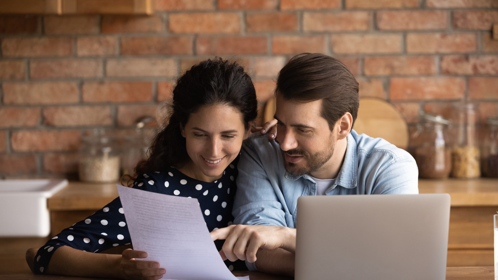 A couple at a kitchen counter reading a printed document together beside an open laptop