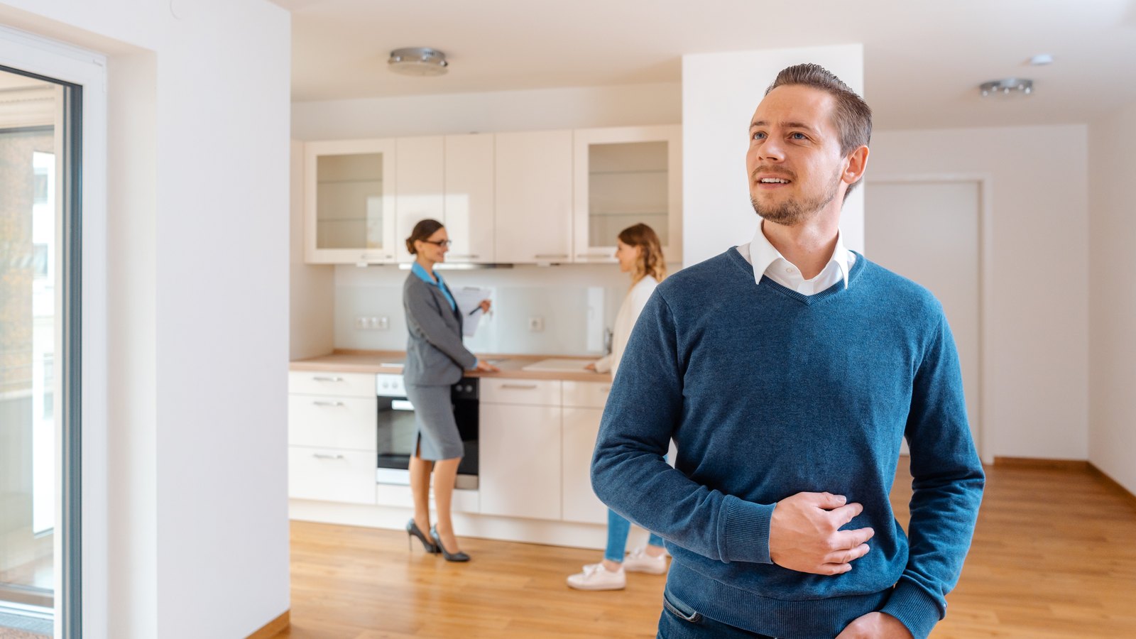 A man standing in an empty apartment during a walkthrough, looking around while two women talk in the kitchen behind him