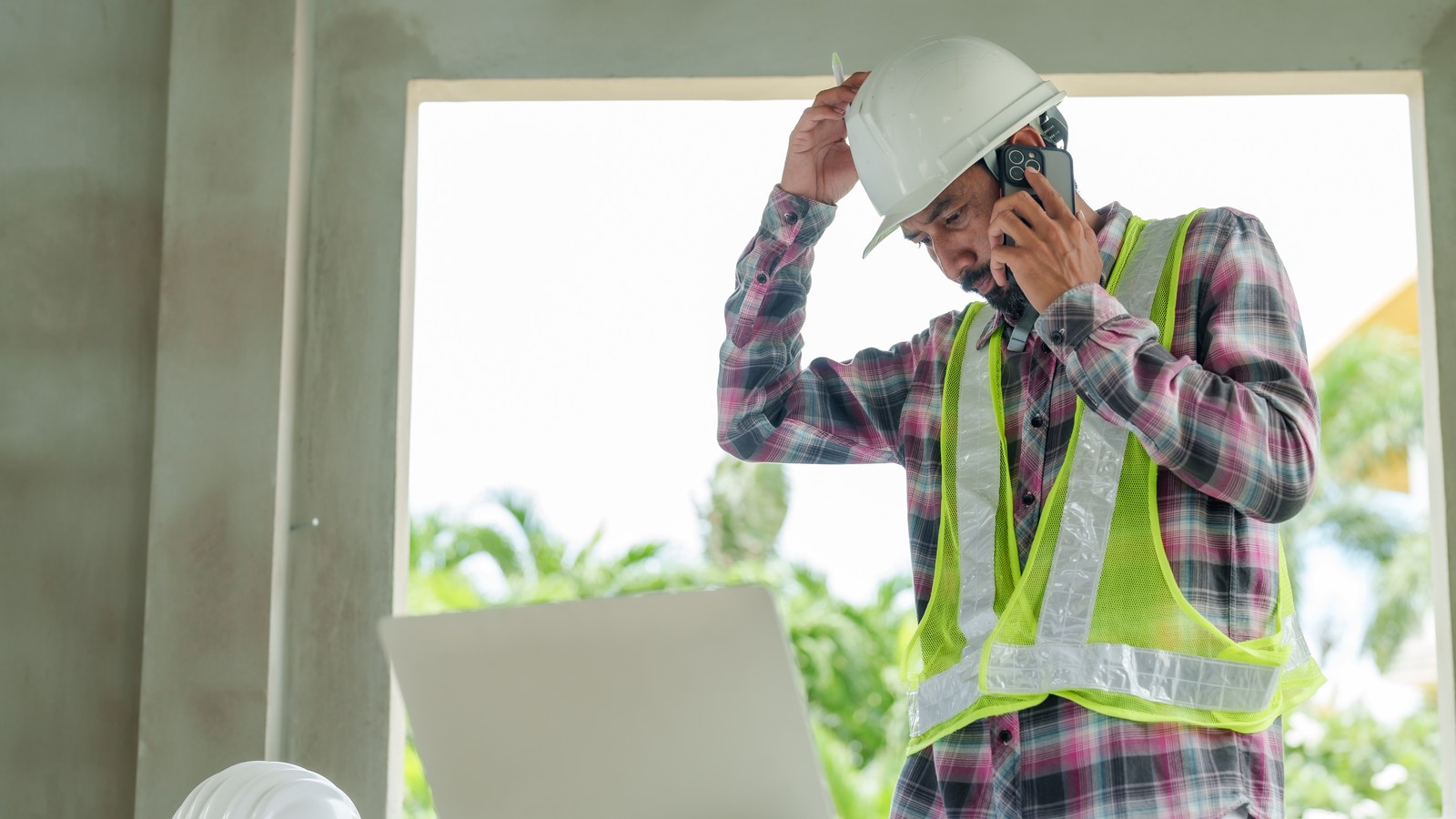 A contractor in a safety vest on the phone inside an unfinished build, hand on his hard hat