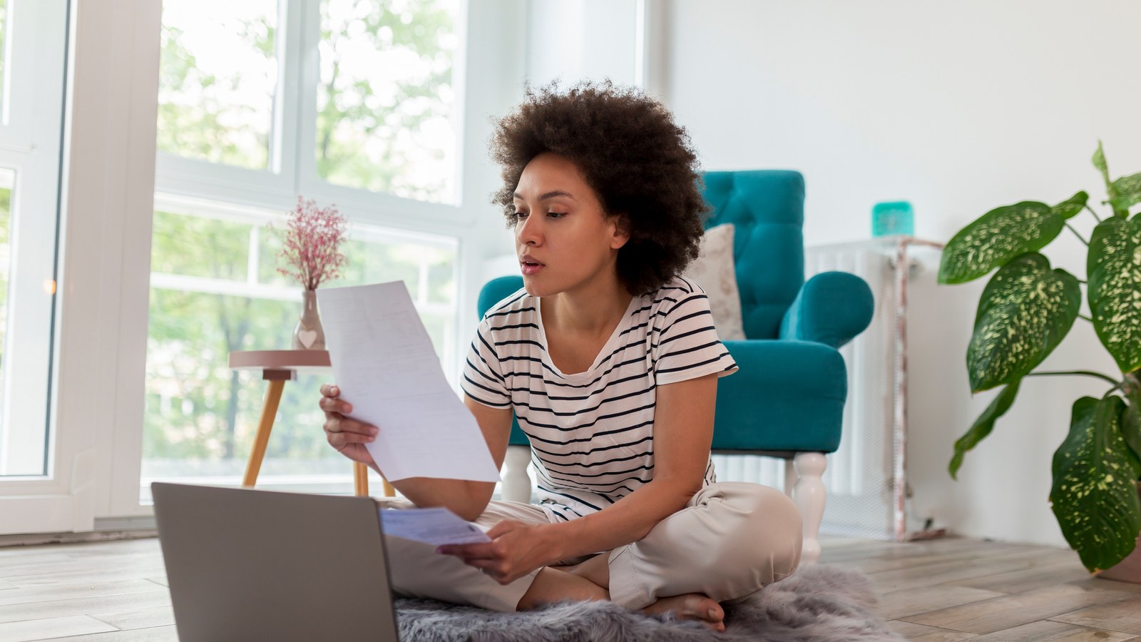 A woman sitting on her living-room floor beside a laptop, reading a printed bill with a concerned look