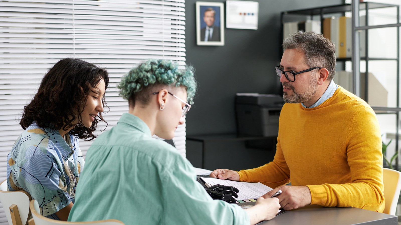 Two people going over paperwork at a table with a man in a yellow sweater who is walking them through it