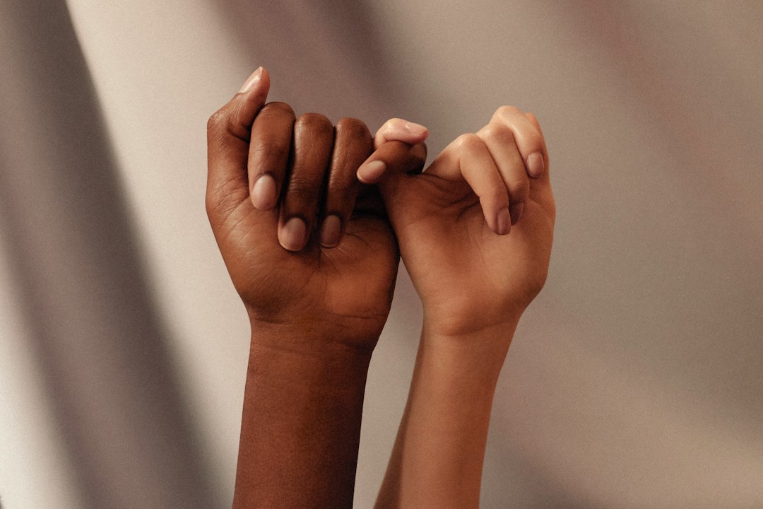 Two raised hands of different skin tones linking little fingers against a soft beige draped background.