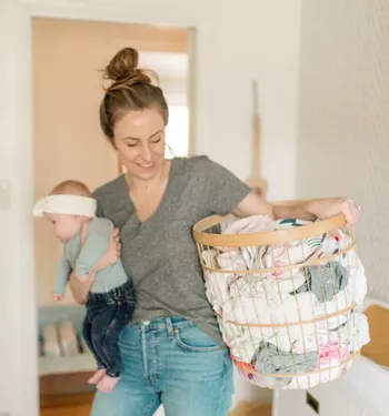 Woman holding a baby in one arm and a laundry basket in the other, smiling while looking down