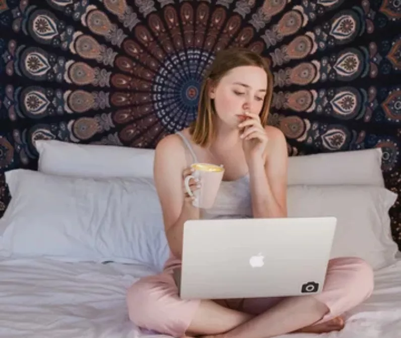 Woman sitting on a bed with a laptop and coffee, mandala tapestry hanging behind her