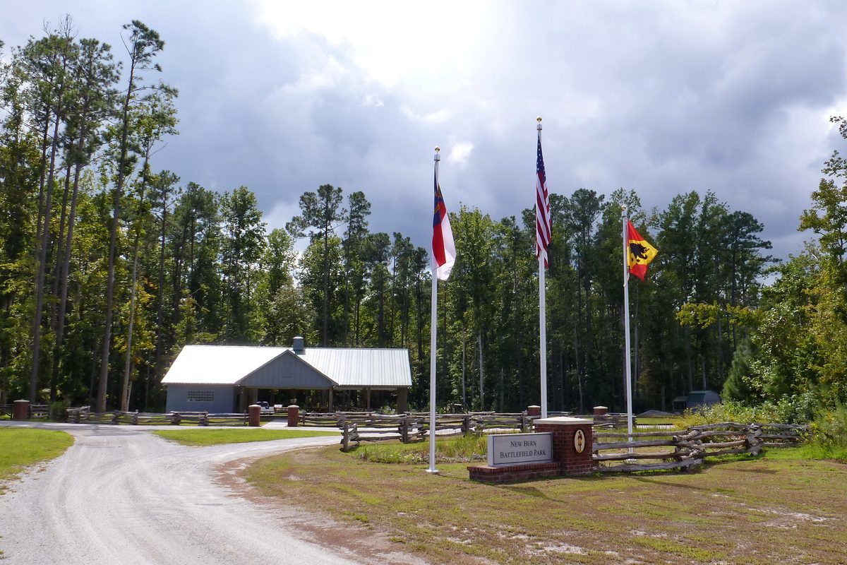 The entrance to New Bern Battlefield Park: a brick sign, three flags and a pavilion among tall pines.