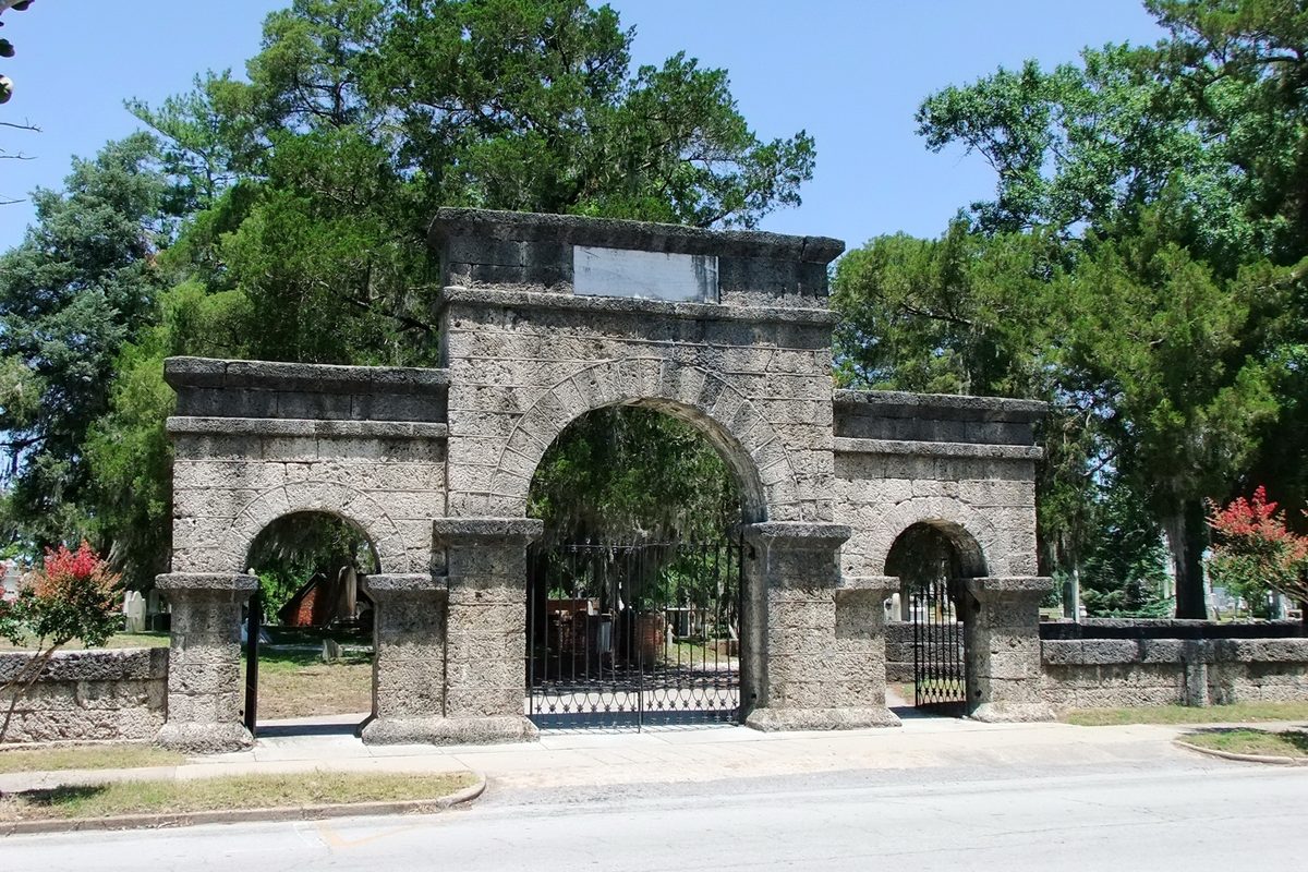The coquina stone entrance arch of Cedar Grove Cemetery, with an iron gate and live oaks behind it.