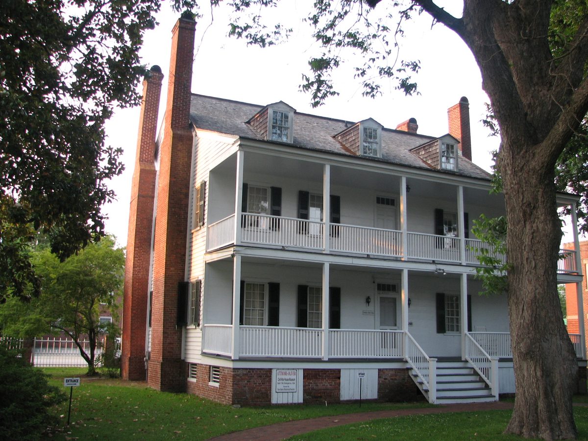 The white Attmore-Oliver House with two-story porches and tall brick chimneys, under an oak tree.