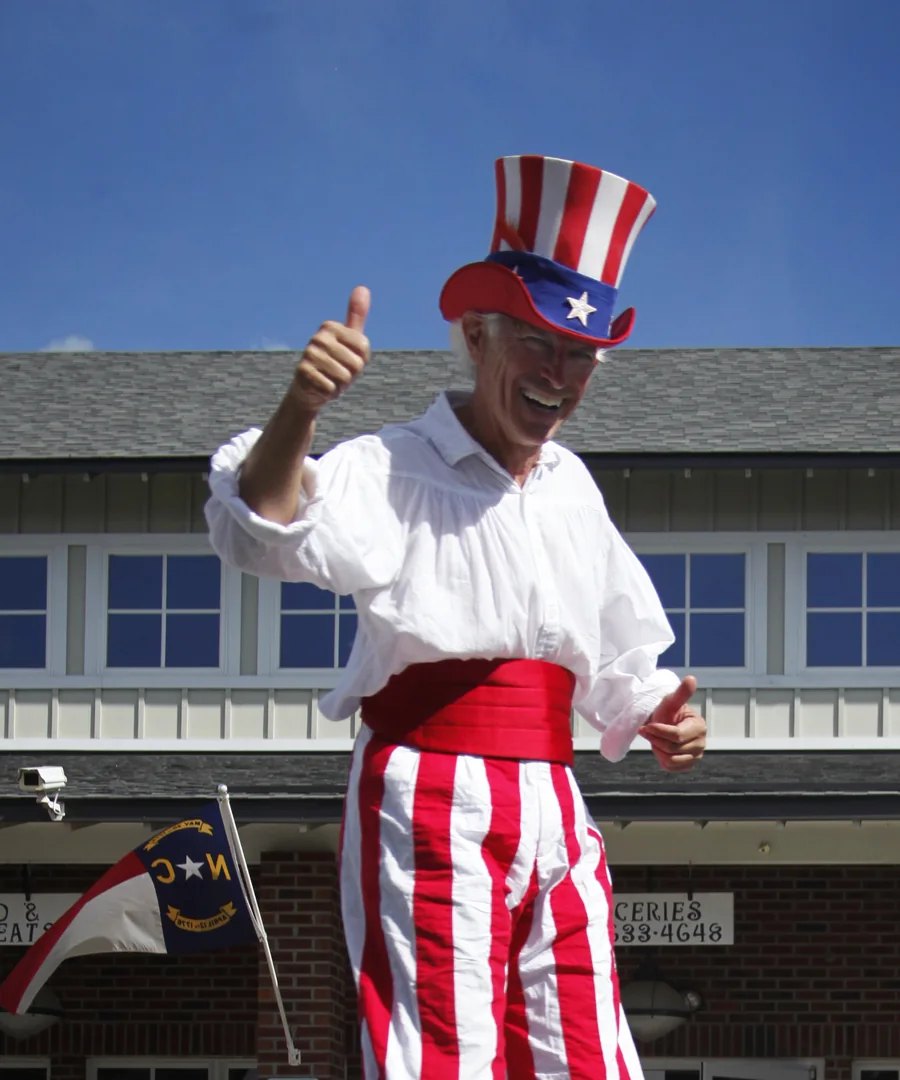 A costumed street performer on stilts in a red, white and blue top hat and striped trousers gives a thumbs up.