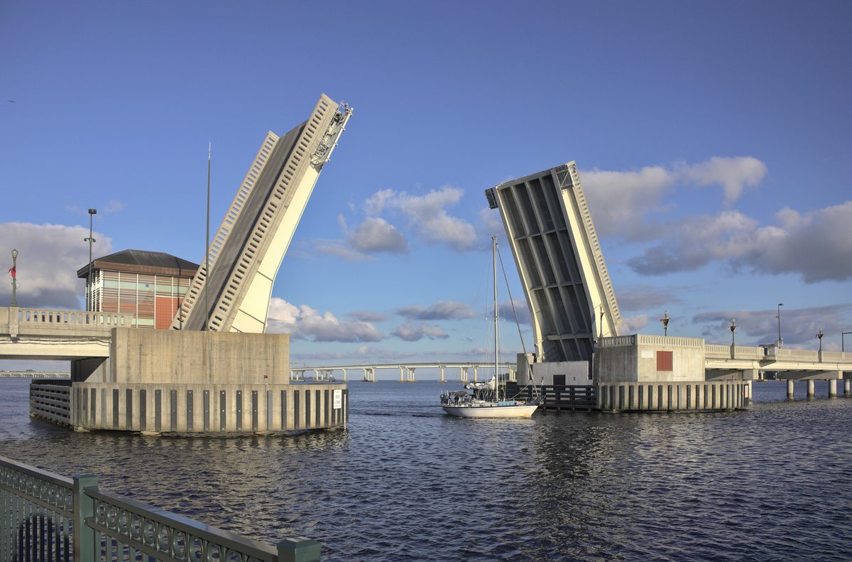 The Riverwalk drawbridge raised over the Neuse River at Union Point Park, with a sailboat passing under a blue sky.