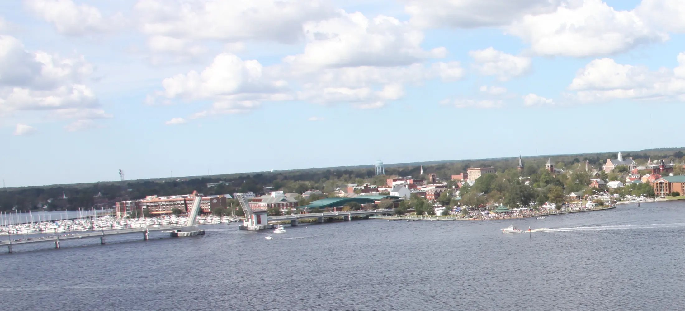 An aerial view across the wide Neuse River to New Bern's downtown waterfront, with the marina at left and the town's rooftops and steeples beyond, under a partly cloudy sky.