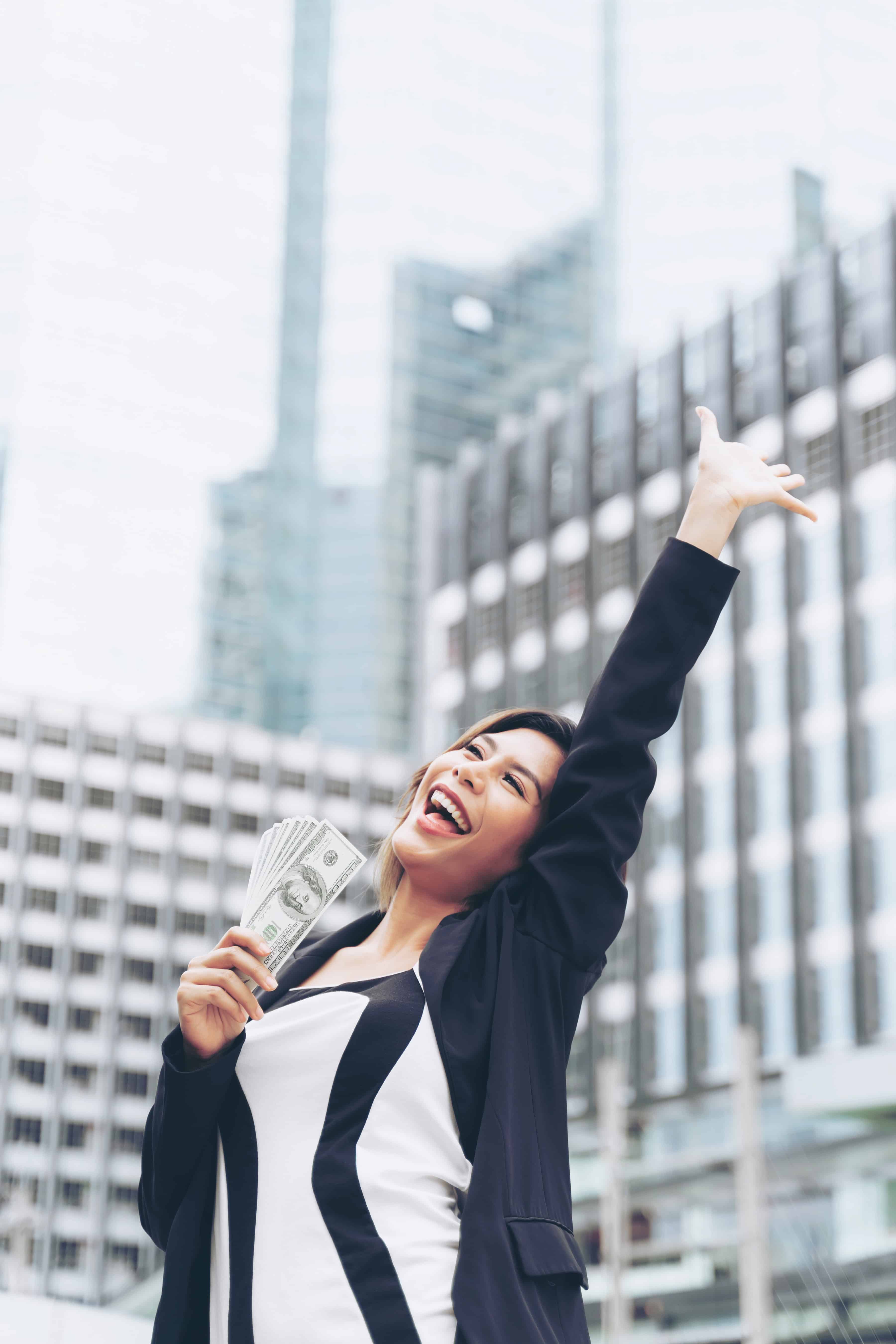 A woman holding a handful of currency smiles and punches the air on a city street surrounded by skyscraper office buildings.