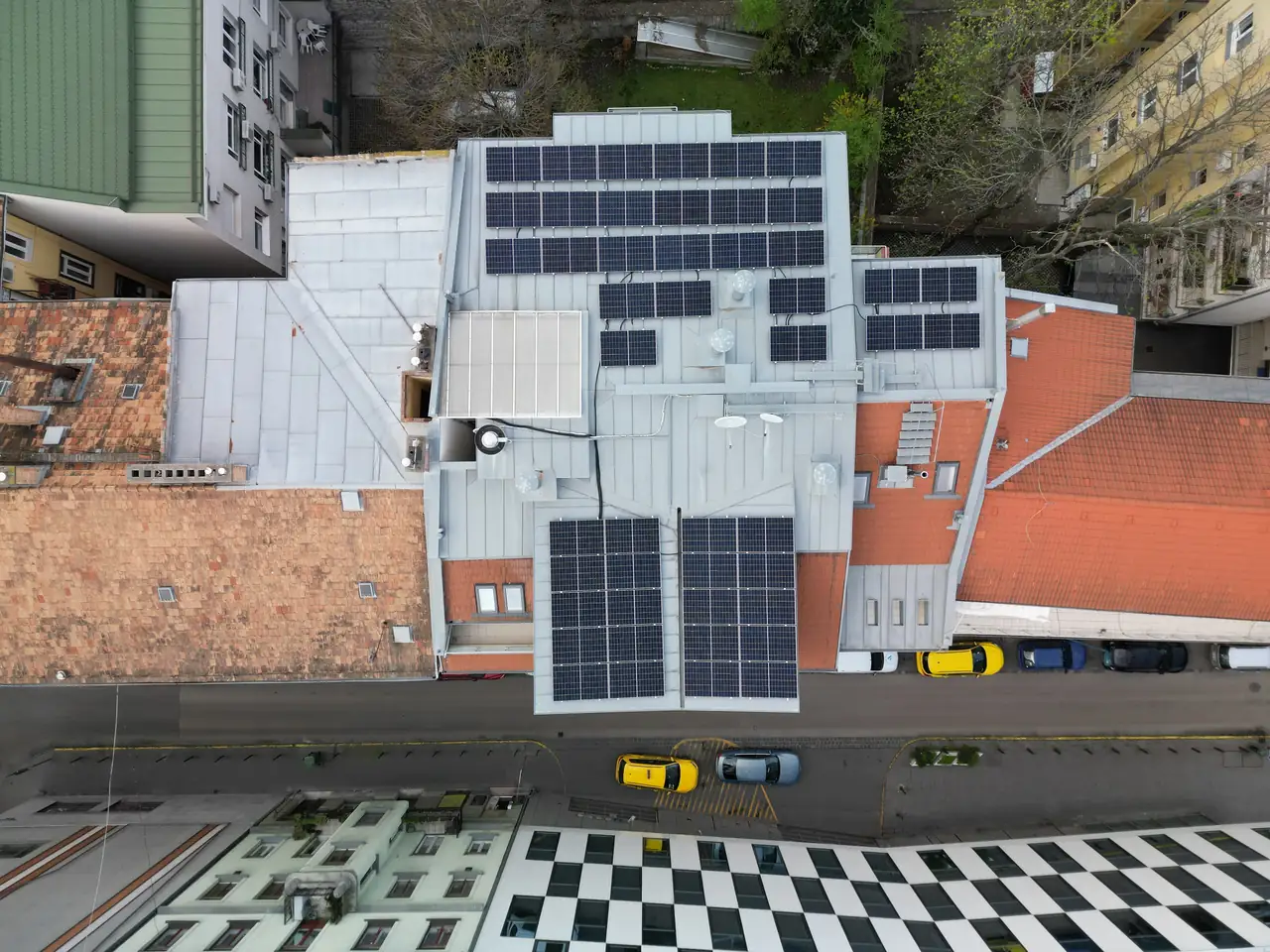 Veszprém városában az Ares Solar Construction Kft. által végzett napelem telepítés látható egy épület tetején. A felvételen jól kivehető, hogy az épület tetőjén több napelem található, amelyek napenergiát hasznosítanak. A környező utcákban parkoló autók és zöld fák is láthatók, ami tovább színesíti a városi tájat.