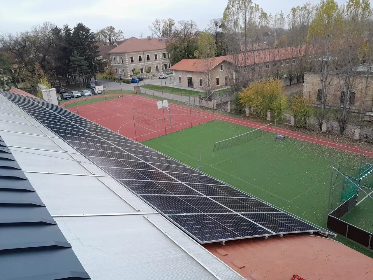 A képen egy Dócon található épület tetején elhelyezett napelemek láthatók, amelyek az Ares Solar Construction Kft. napelem telepítési szolgáltatásának részei. A háttérben sportpályák és egy régi, felújított épület látható, míg a környező fák színes őszi levelekkel díszítik a tájat. Az idő borongós, ami tovább növeli a kép hangulatát.