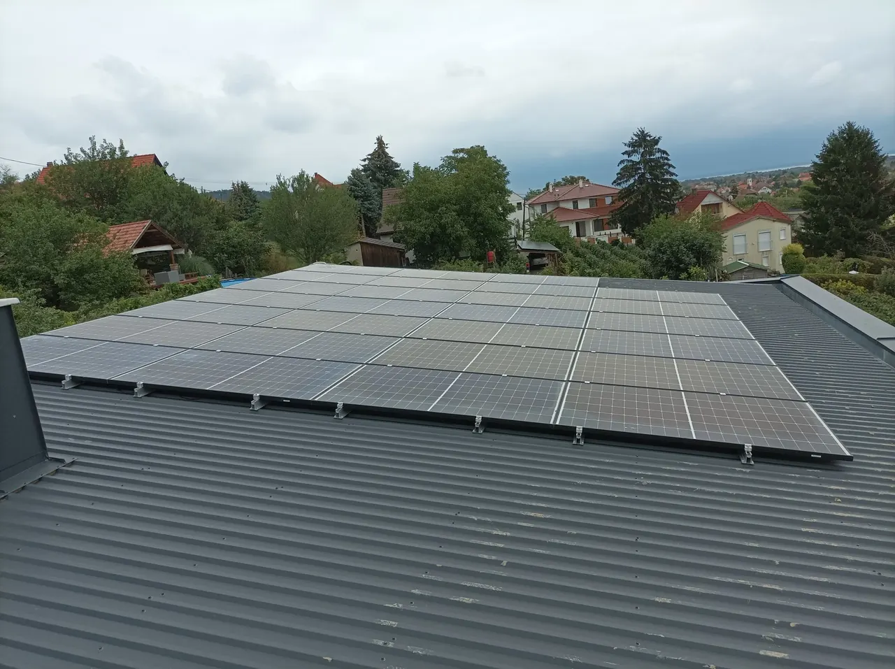A képen jól látható egy Dócon, az Ares Solar Construction Kft. által telepített napelem rendszer, amely egy modern, szürke tetőn helyezkedik el. A napelemek szépen sorakoznak egymás mellett, miközben a háttérben zöld fák és lakóépületek láthatók. Ez a napelem telepítés a fenntartható energiaforrások elősegítését szolgálja, bemutatva a cég szakértelmét a napelem telepítés terén.