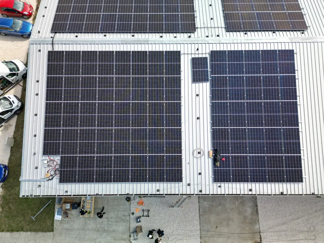 A képen a Dócon található Ares Solar Construction Kft. napelem telepítési munkálatai láthatók. A tetőn elhelyezett napelemek sorakoznak, amelyek napfényt gyűjtenek a zöld energia előállítása érdekében. Az épület környezetében néhány munkaeszköz és autó is megfigyelhető, ami a telepítési folyamatot jelzi.