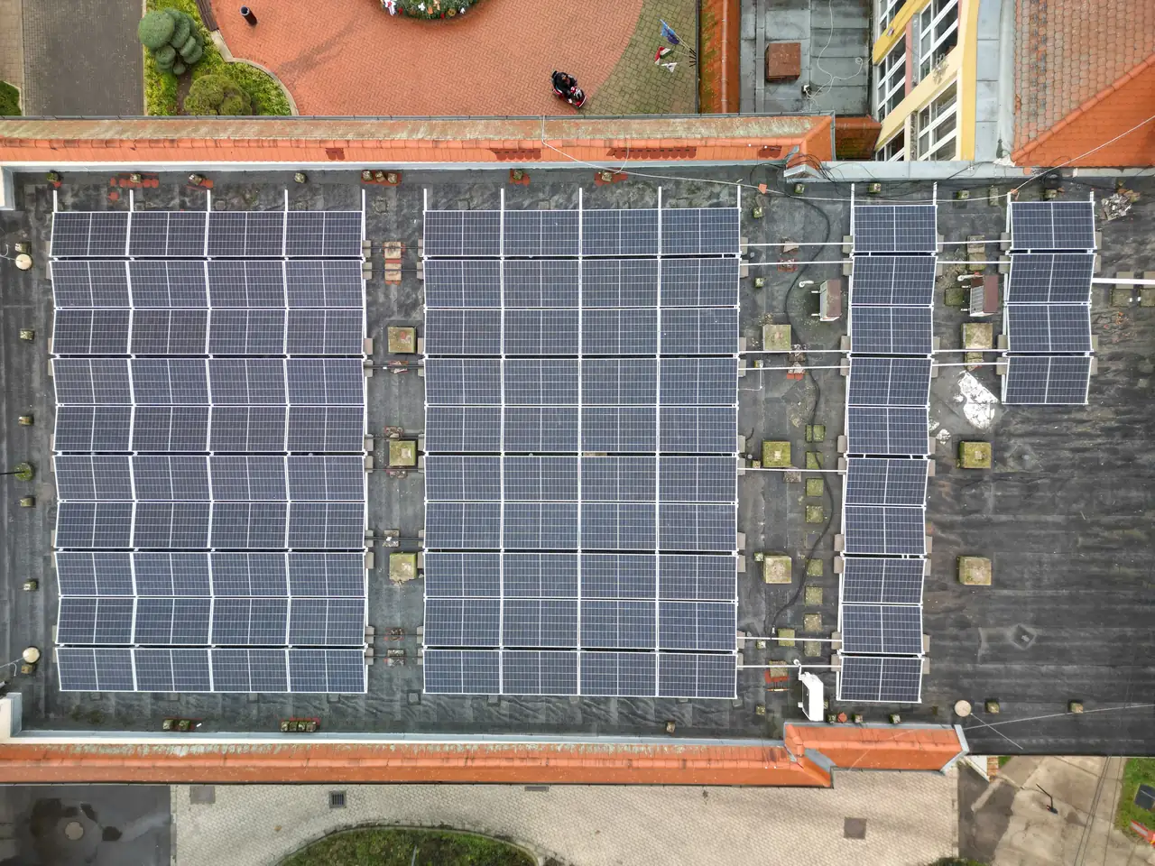 A képen Dóc városában található épület tetején elhelyezett napelemek láthatók, amelyek az Ares Solar Construction Kft. Napelem Telepítés szolgáltatásának eredményeként kerültek kihelyezésre. Az átlátható, modern napelem rendszer hatékony energiafelhasználást biztosít, és a tető egyenletesen van borítva a napelemekkel. A háttérben zöld területek és más épületek is láthatók.