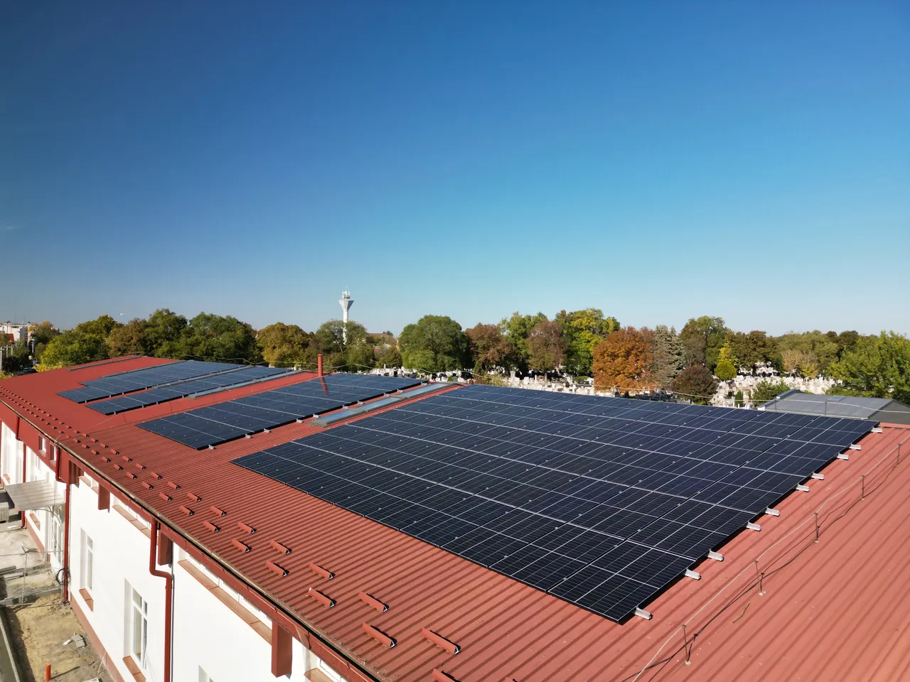 Békéscsabán, az Ares Solar Construction Kft. szakemberei napelemeket telepítenek egy modern ház tetejére. A képen jól látható a fényes, fehér fémlemez tető, amelyen a napelemek helyezkednek el. A zöldellő fák és a környezet nyújtotta háttér hangsúlyozza a fenntartható energiaforrások iránti elköteleződést a napelem telepítés terén.