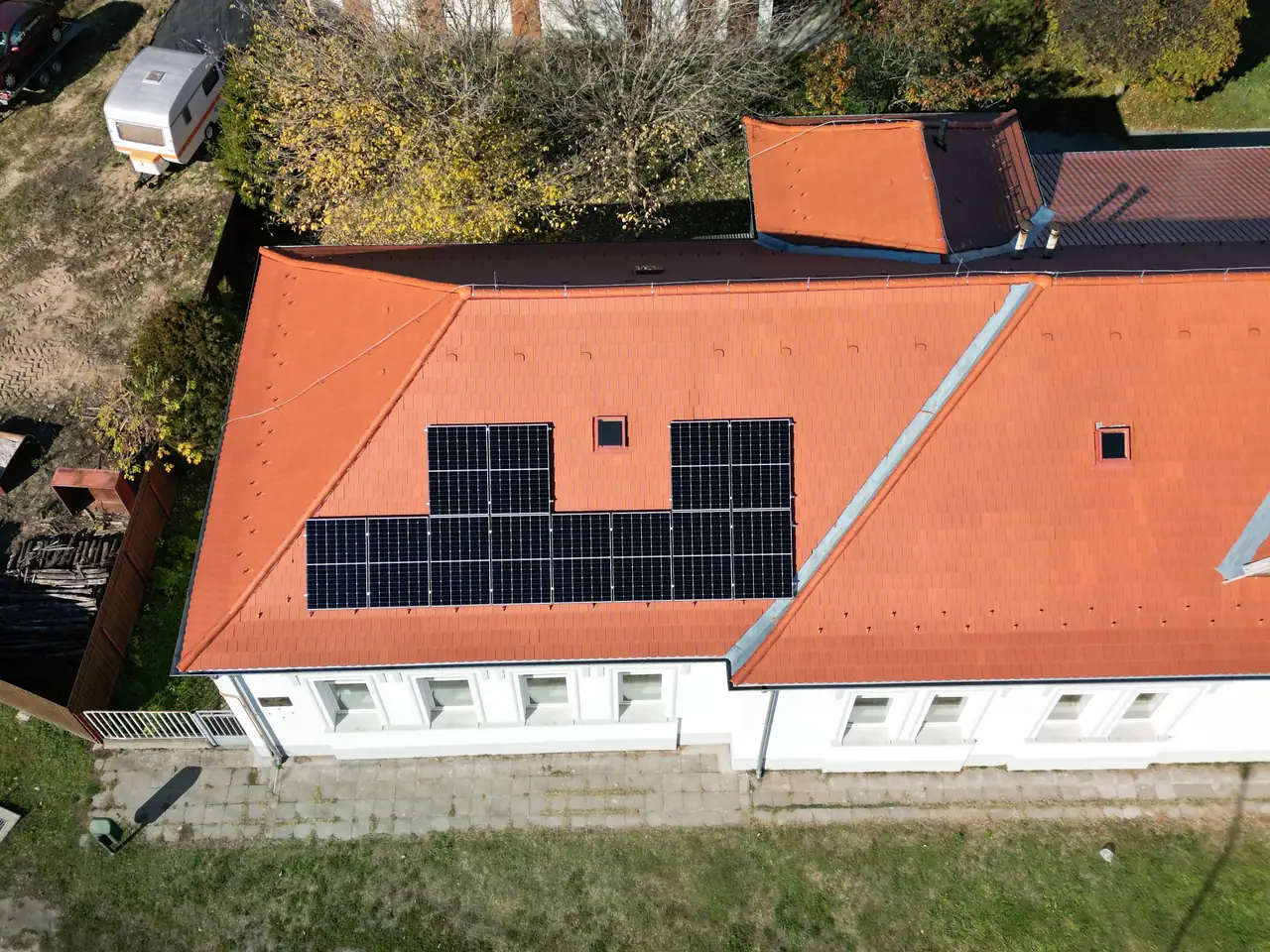 A képen egy Dócon található ház látható, amelynek tetőjén napelemek vannak telepítve. A napelemek szimmetrikusan helyezkednek el a piros cseréptetőn, amely jól illik a környező zöld fűhöz és a fákhoz. Az Ares Solar Construction Kft. napelem telepítési szolgáltatása jól láthatóan hozzájárul a környezetbarát energiahasználathoz.