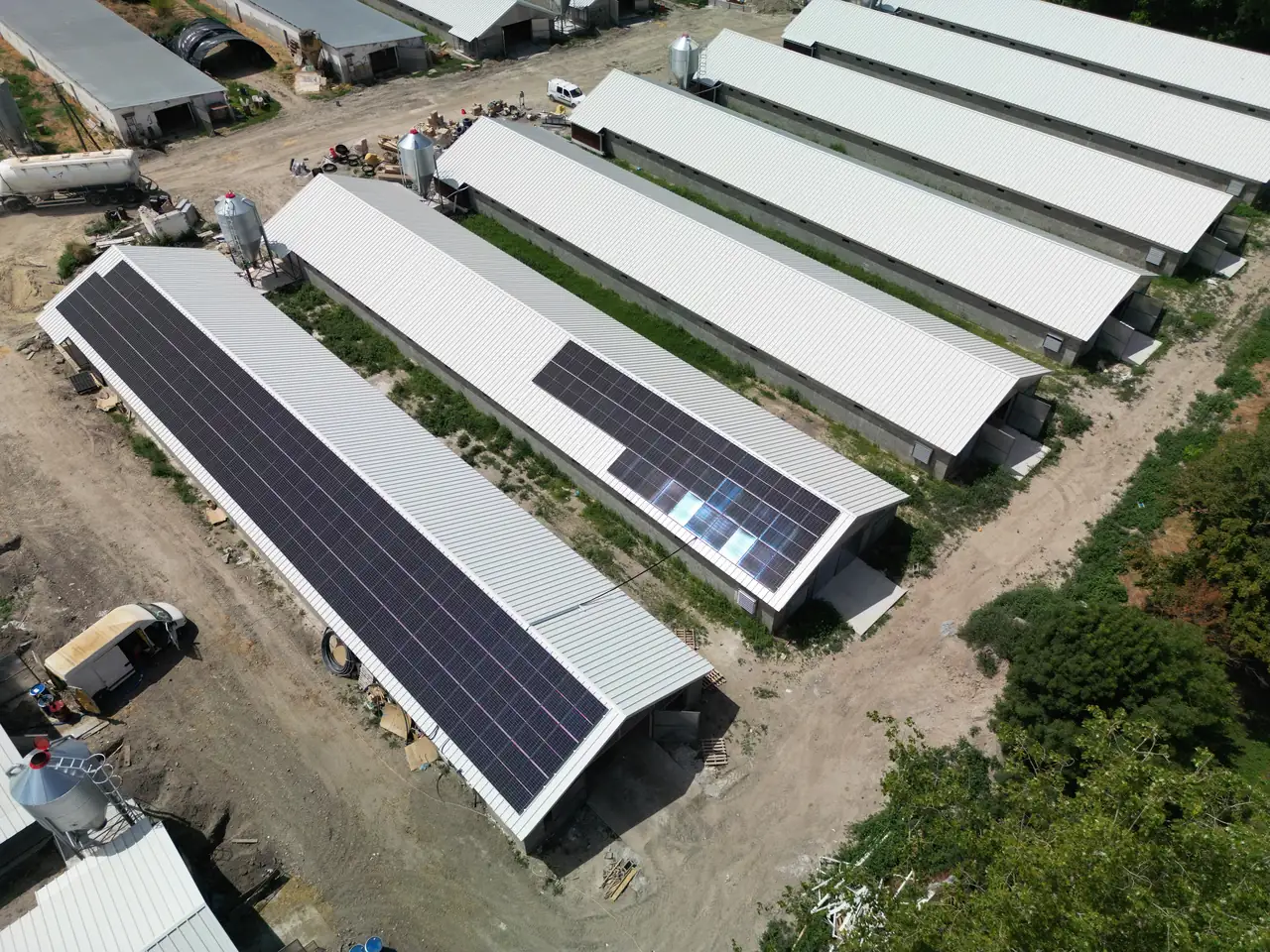 A képen Dócon található napelem telepítés látható, ahol az Ares Solar Construction Kft. által telepített napelemek egy mezőgazdasági épület tetején helyezkednek el. Az épületek fehér fém tetői jól láthatóak, míg a napelemek sötét színe kontrasztot ad a világos háttérnek. A környező területen zöld növényzet és egyéb épületek is láthatók, amelyek a telepítés helyszínét alkotják.