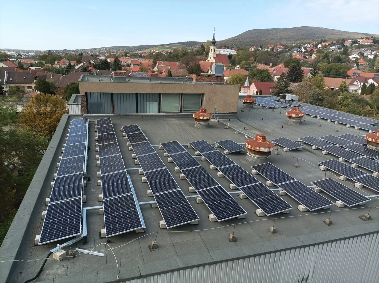 Dóc városában, az Ares Solar Construction Kft. napelem telepítési projektje látható, ahol egy modern épület tetején több sor napelem található. A képen a napelemek szakszerű elhelyezése és a városi táj részletei is jól látszanak, bemutatva a fenntartható energia használatát a helyi közösségben. A háttérben a település jellegzetes épületei és a dombos táj is felsejlik.