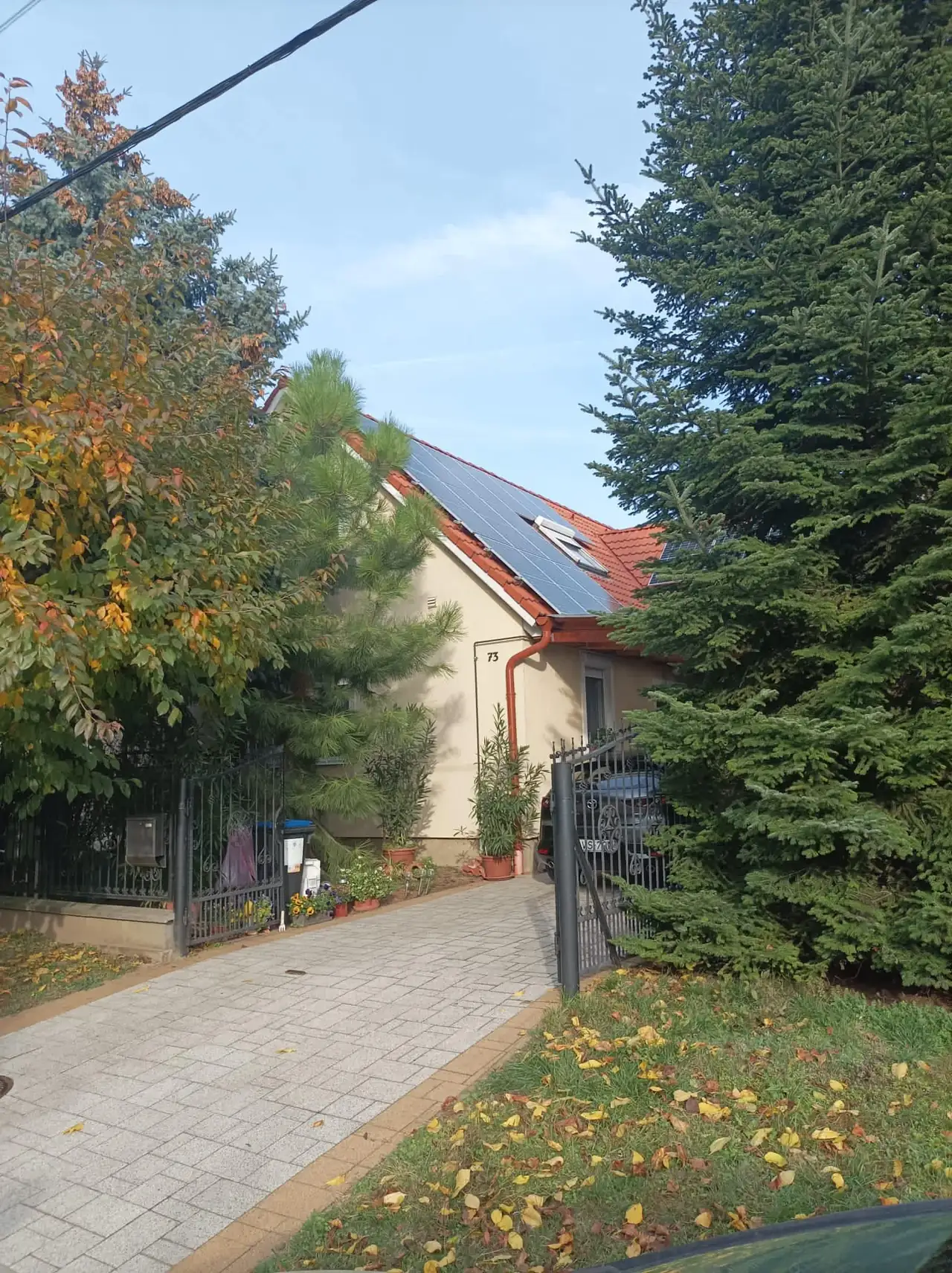 Balaton városában, az Ares Solar Construction Kft. napelem telepítési szolgáltatásának bemutatása látható. A képen egy ház látható, amelynek tetején napelemek találhatók, körülötte gyönyörű zöld növényzet és fák. Az épület előtt rendezett járda és kerítés áll, amely barátságos hangulatot áraszt. Az őszi falevelek színei még inkább kiemelik a környezet szépségét.