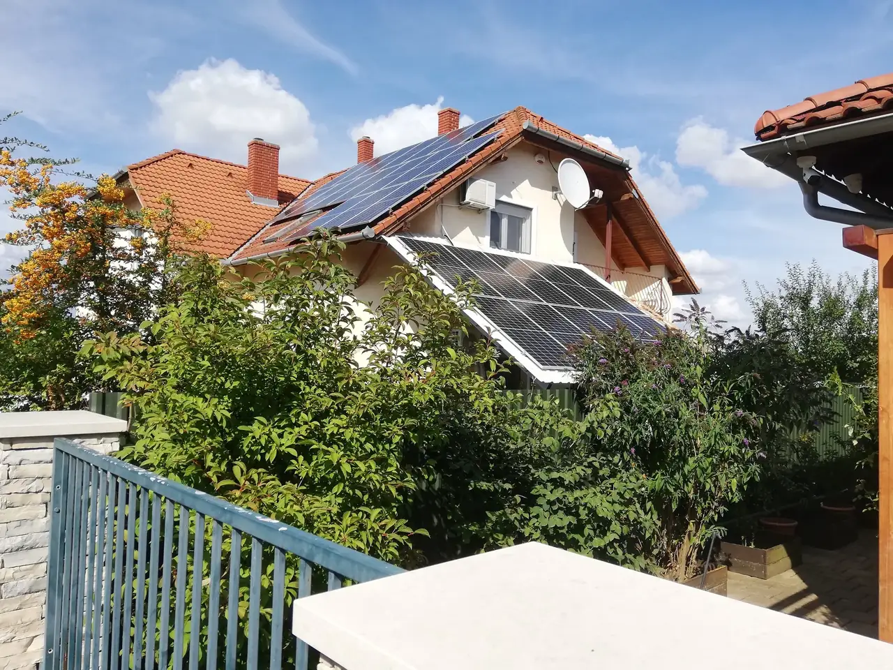 A képen egy családi ház látható, amelynek tetején napelemek vannak telepítve. A ház körül buja zöld növényzet és kerítés található, ami kellemes, természet közeli környezetet teremt. A napelem telepítés részletei jól láthatóak, ami a fenntartható energiahasználat fontosságát hangsúlyozza. Az Ares Solar Construction Kft. szakemberei által végzett napelem telepítés {city} területén hozzájárul a környezetbarát energiaforrások kihasználásához.