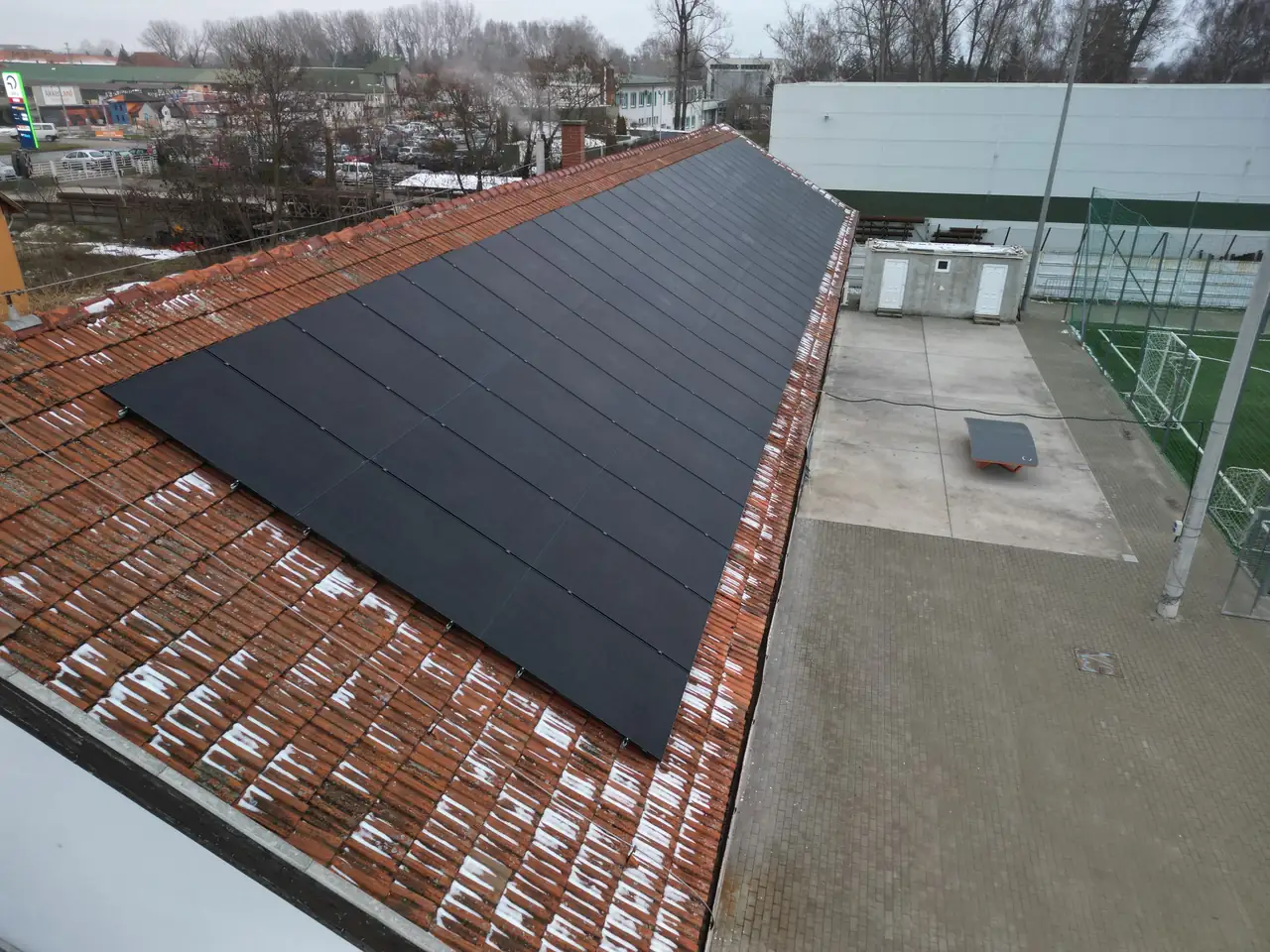 A képen egy Szolnokon található épület tetője látható, amelyet Ares Solar Construction Kft. napelemekkel szerelt fel. A tetőn egy sor fekete napelem található, amelyek hatékonyan használják a napenergiát. Az épület környezetében láthatóak más épületek és egy aszfaltozott terület is, amely a napelem telepítésének helyszínét mutatja.