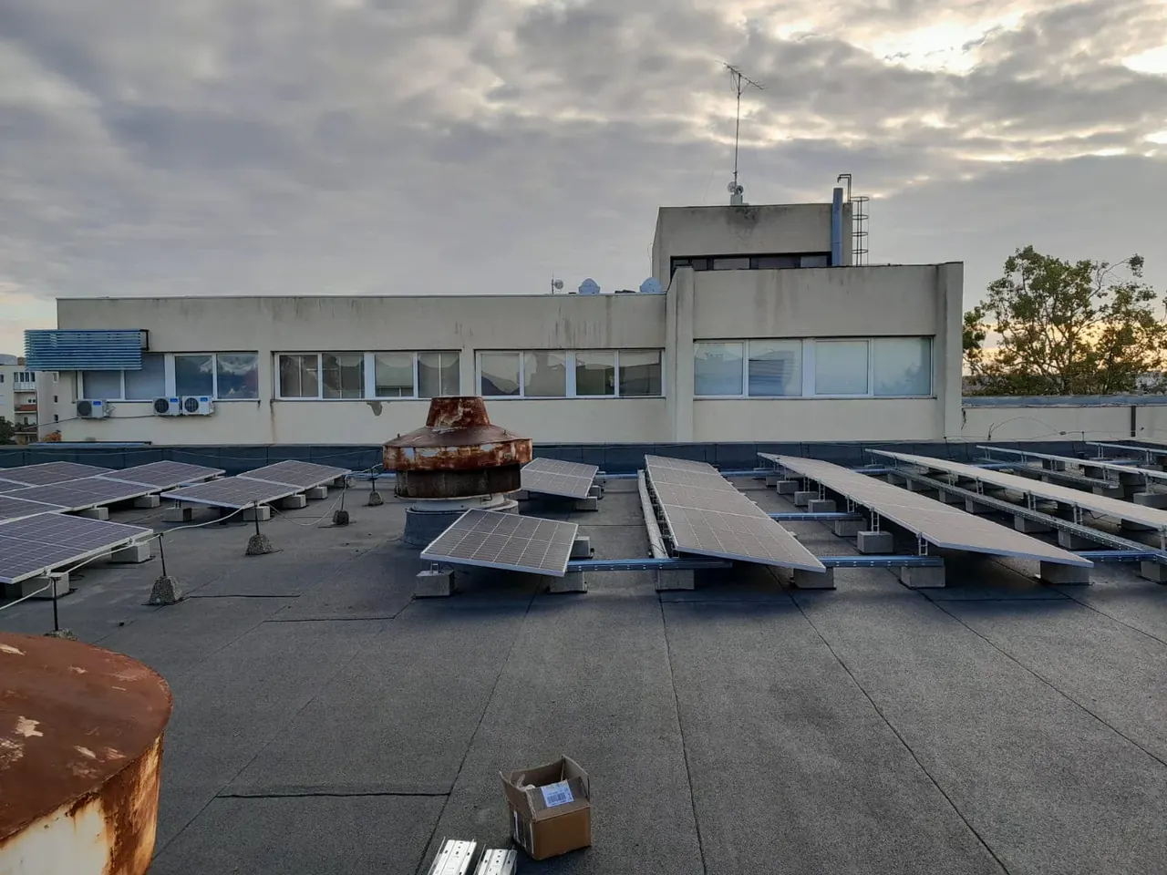 Napelemek sorakoznak a Dócon található épület tetején, amely a Ares Solar Construction Kft. által végzett napelem telepítést mutatja be. A tetőn láthatóak a napenergia hasznosítására szolgáló panelek, valamint a környező városi táj, amely a modern megújuló energiaforrások iránti elkötelezettséget tükrözi. A háttérben a felhős égbolt és a városi épületek láthatók.