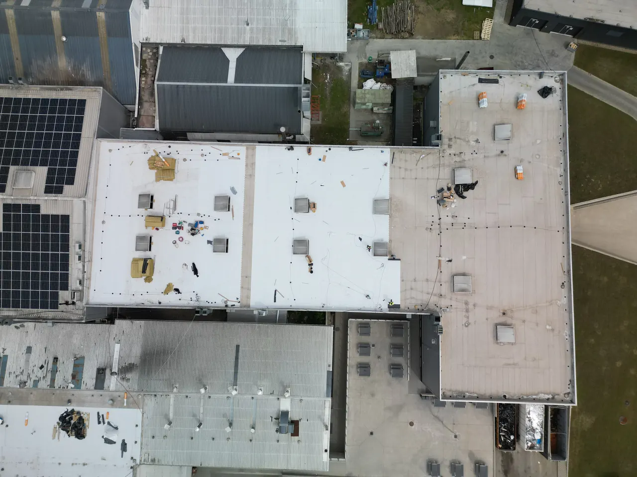 Dóc városában, az Ares Solar Construction Kft. napelem telepítésének folyamatát mutatja az a légi felvétel, amelyen egy nagyobb ipari épület tetője látható. A képen jól láthatók a napelemek és a tetőn elhelyezett különböző szerelési anyagok, valamint a munkálatokat végző szakemberek. A környező épületek is részben láthatók, amelyek a telepítés helyszínét ölelik körül.