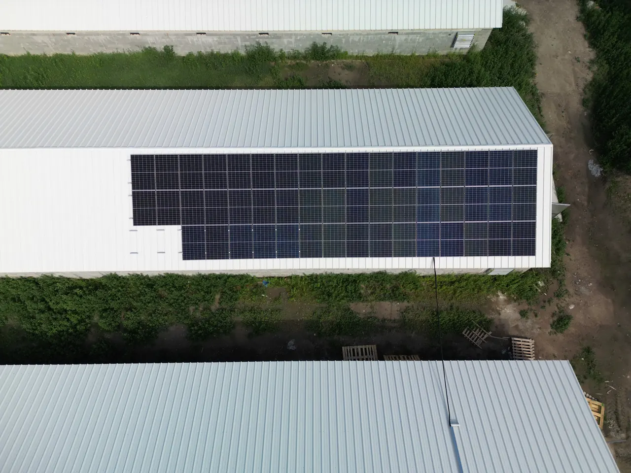 Eger városában található Ares Solar Construction Kft. napelem telepítési projektje látható, ahol egy ipari épület tetején elhelyezett napelem panelek sorakoznak. A tető fehér színű, és a zöld növényzet körbeveszi az épületet. A napenergia kihasználása és a fenntarthatóság érdekében telepített napelemek modern megoldást kínálnak az energiafogyasztás csökkentésére.