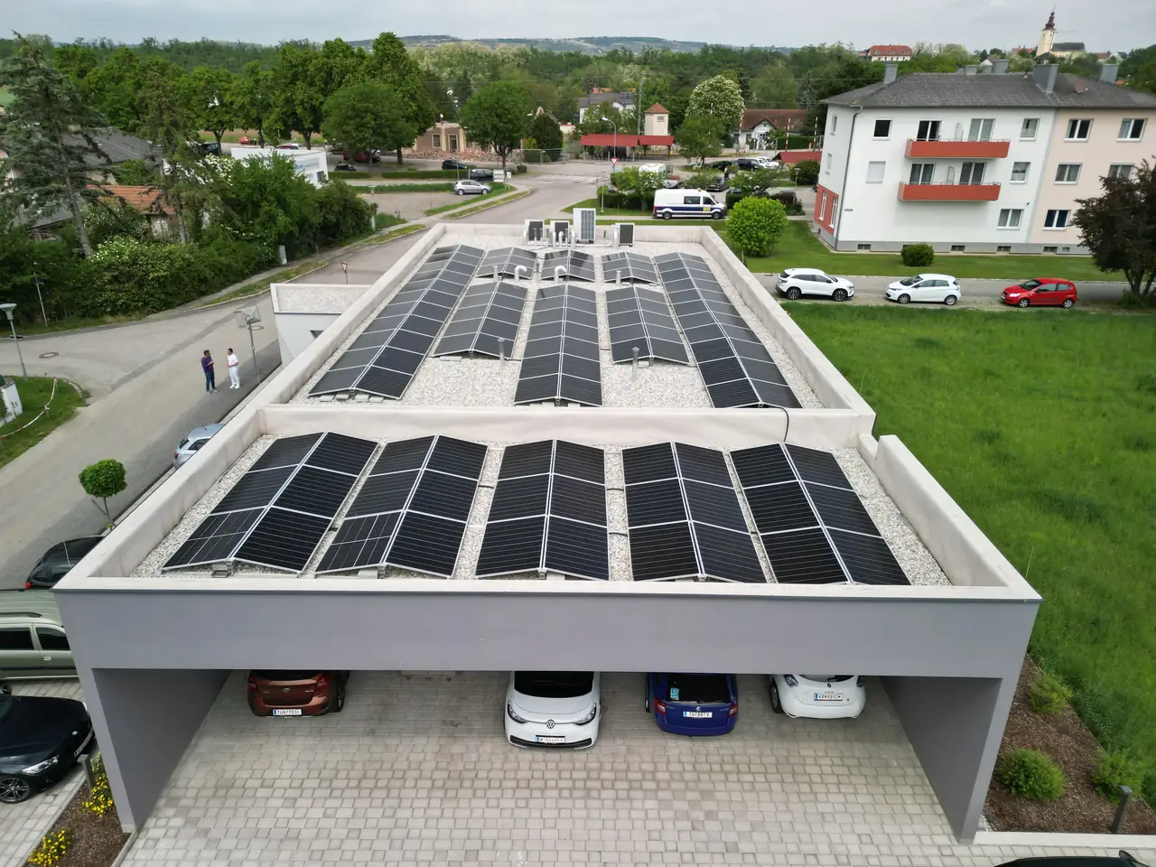 Napelemekkel felszerelt modern épület tetője látható Dócon, ahol az Ares Solar Construction Kft. napelem telepítési szolgáltatását nyújtja. Az épület alatt parkoló autók sorakoznak, míg a zöld területen fákkal szegélyezett udvar található. Az okosotthonok kivitelezésére specializálódott cég munkájának köszönhetően a tetőn elhelyezett napelemek hatékonyan termelik az energiát.