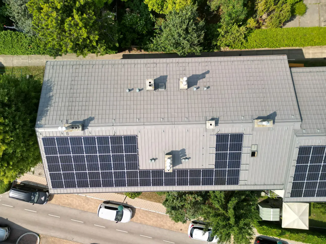 A képen egy budapesti épület teteje látható, amelyen napelemek sorakoznak, jelezve az Ares Solar Construction Kft. napelem telepítési tevékenységét. Az épület körül zöld növényzet található, míg az utcán parkoló autók láthatók. A napelemek hatékonyan hasznosítják a napenergiát, hozzájárulva a fenntartható energiahasználathoz Budapest területén.