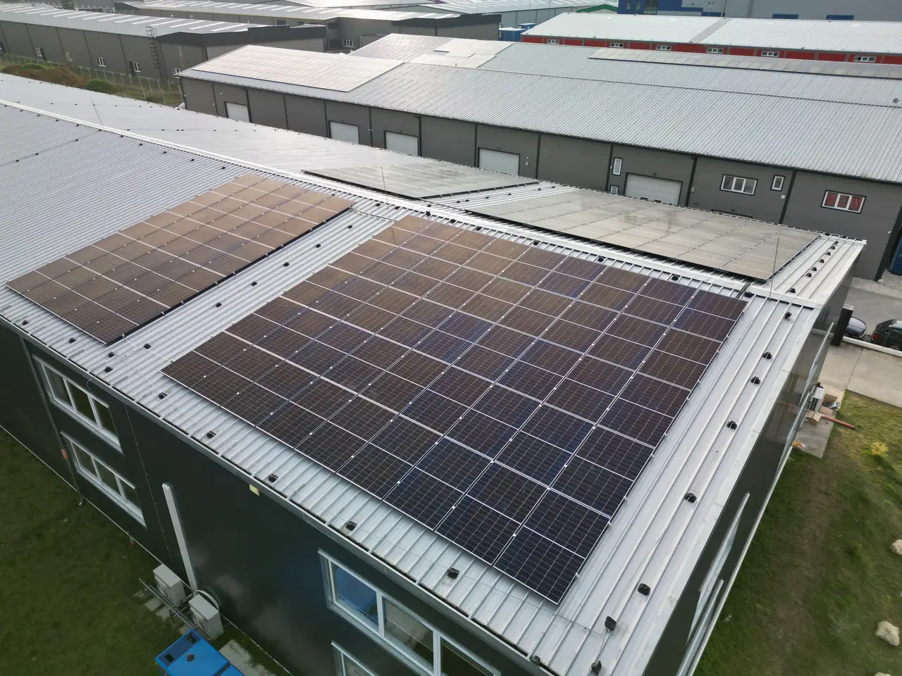 Dunaújvárosban, az Ares Solar Construction Kft. által végzett napelem telepítés látható egy ipari épület tetején. A képen jól látszanak a fekete napelemek, amelyek a tető síkján helyezkednek el, és a környező épületek is részben láthatók. A napfényben csillogó napelemek modern technológiát képviselnek, hozzájárulva a fenntartható energiahasználathoz.